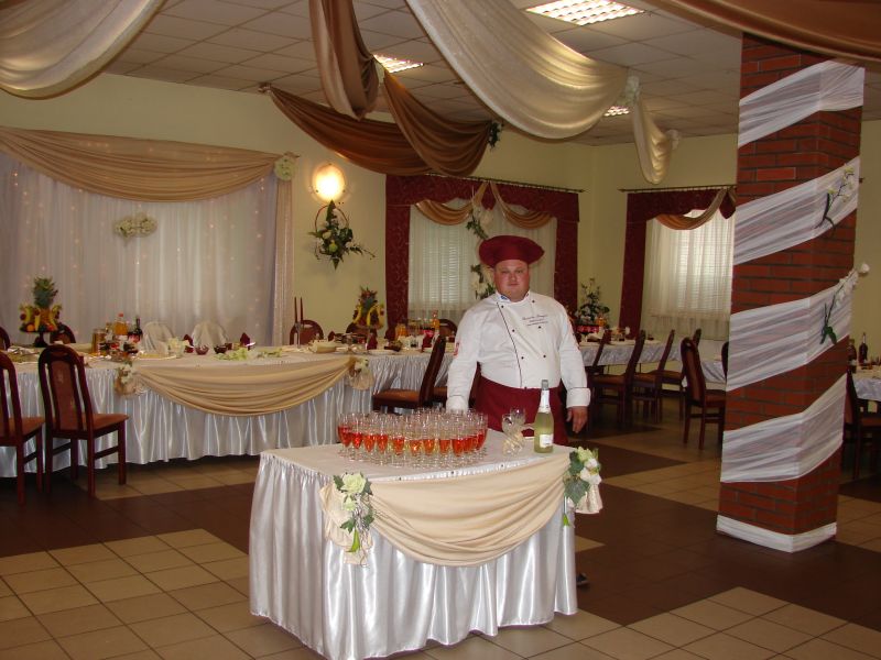 Sala bankietowa z dekoracjami, nakrytymi stołami i bufetem z napojami, kucharz w stroju galowym na pierwszym planie.